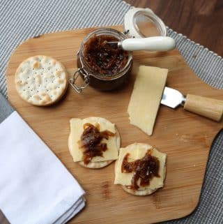 Caramelised onion chutney in a jar on a wooden board with water biscuits, cheese, cheese knife, butter knife and napkin