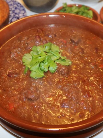 Brisket chilli in a large terracotta serving dish with cornbread, rice, cheese and chopped chillis in the background