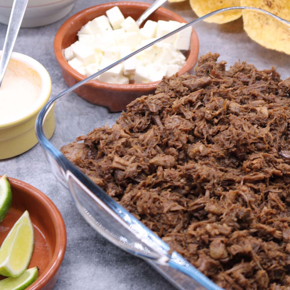 Barbacoa pulled beef in square serving dish with tacos, cheese and limes in background