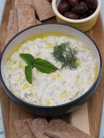 Feta and charred spring onion tzatziki in bowl with mint and dill, sitting on wooden board with olives and pita bread