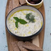 Feta and charred spring onion tzatziki in bowl with mint and dill, sitting on wooden board with olives and pita bread