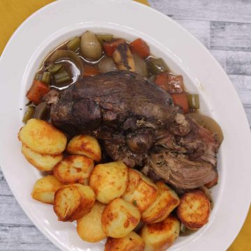 Slow cooker lamb shoulder on white oval serving dish with roast potatoes and vegetables