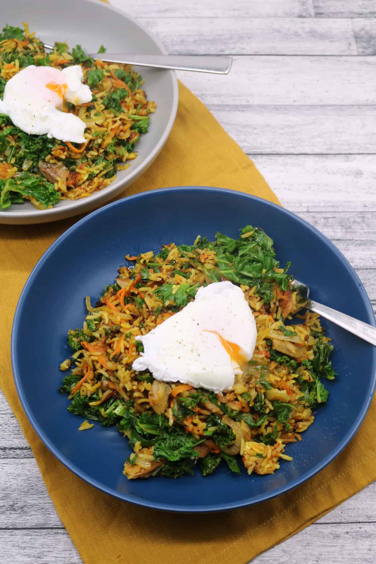 Spiced kipper fried rice topped with poached egg in blue and grey bowls sitting on yellow napkin with fork