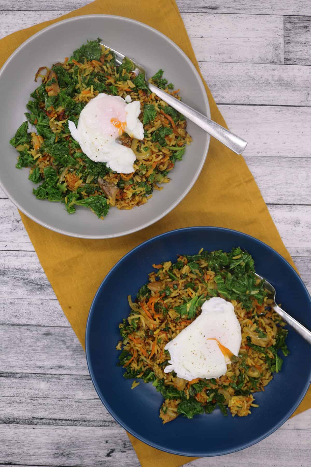 Spiced kipper fried rice topped with poached egg in blue and grey bowls sitting on yellow napkin with fork