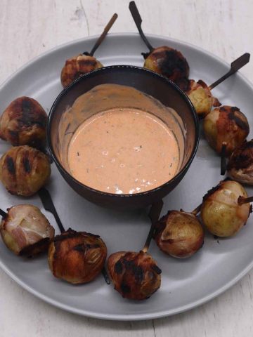 Baby potatoes wrapped in bacon with skewers alongside bowl of Harissa cheese dip