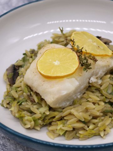 Baked cod with creamy mushroom and leek orzo in bowl topped with lemon slices and thyme