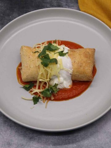 Chicken chimichanga on grey plate covered in cheese, coriander and soured cream on top of tomato sauce