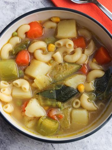 chunky vegetable and pasta soup in a bowl with a spoon and orange napkin