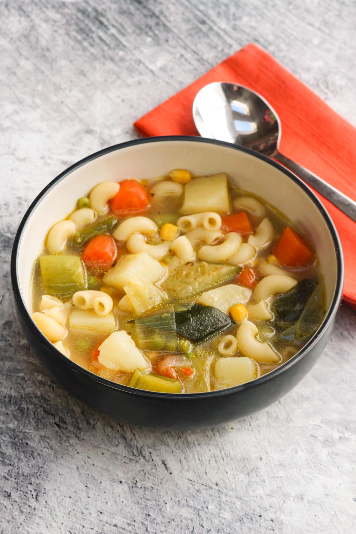 chunky vegetable and pasta soup in a bowl with a spoon and orange napkin