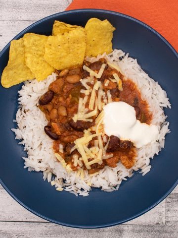 smoky pork and beans in a blue bowl with rice, nachos and soured cream next to orange napkin