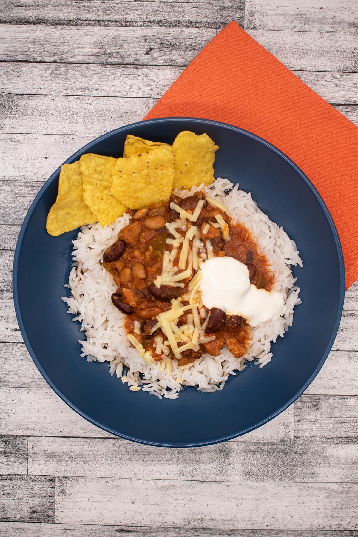 smoky pork and beans in a blue bowl with rice, nachos and soured cream next to orange napkin