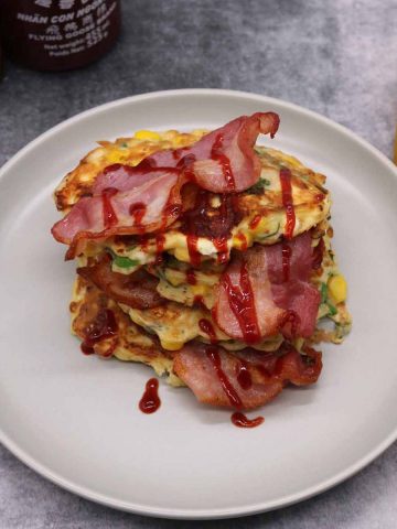 Sweetcorn Feta and Spring onion pancakes stacked on a grey plate with bacon and drizzle of Sriracha