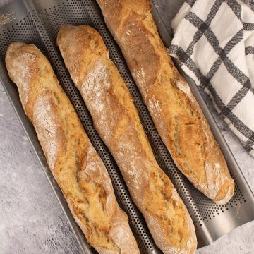 Three baguettes in a baguette baking tray beside black and white towel