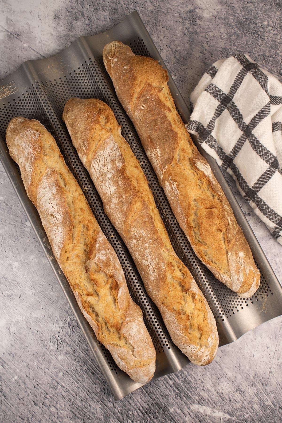 Three baguettes in a baguette baking tray beside black and white towel