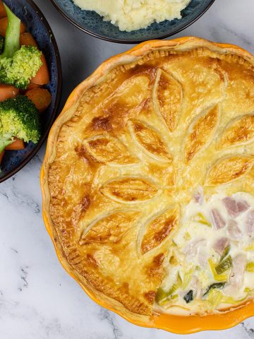 Creamy chicken, gammon and leek pie in a round pie dish with bowls of veg and mash in the background