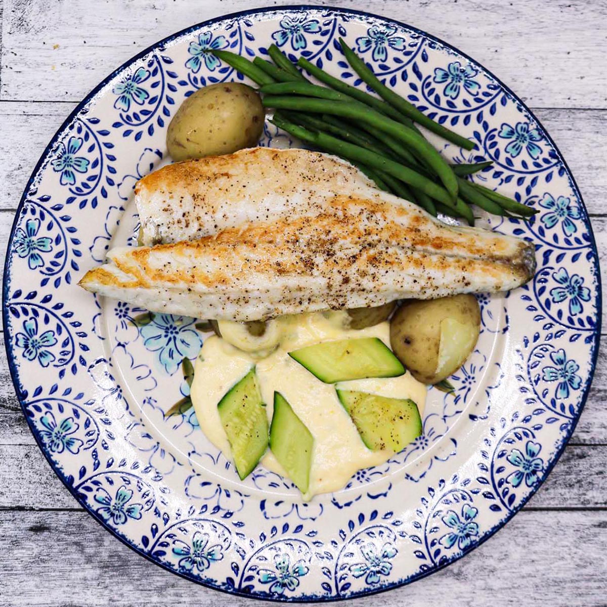Sea Bass fillet on a blue and white plate with lemony sauce, boiled new potatoes, green beans and courgette