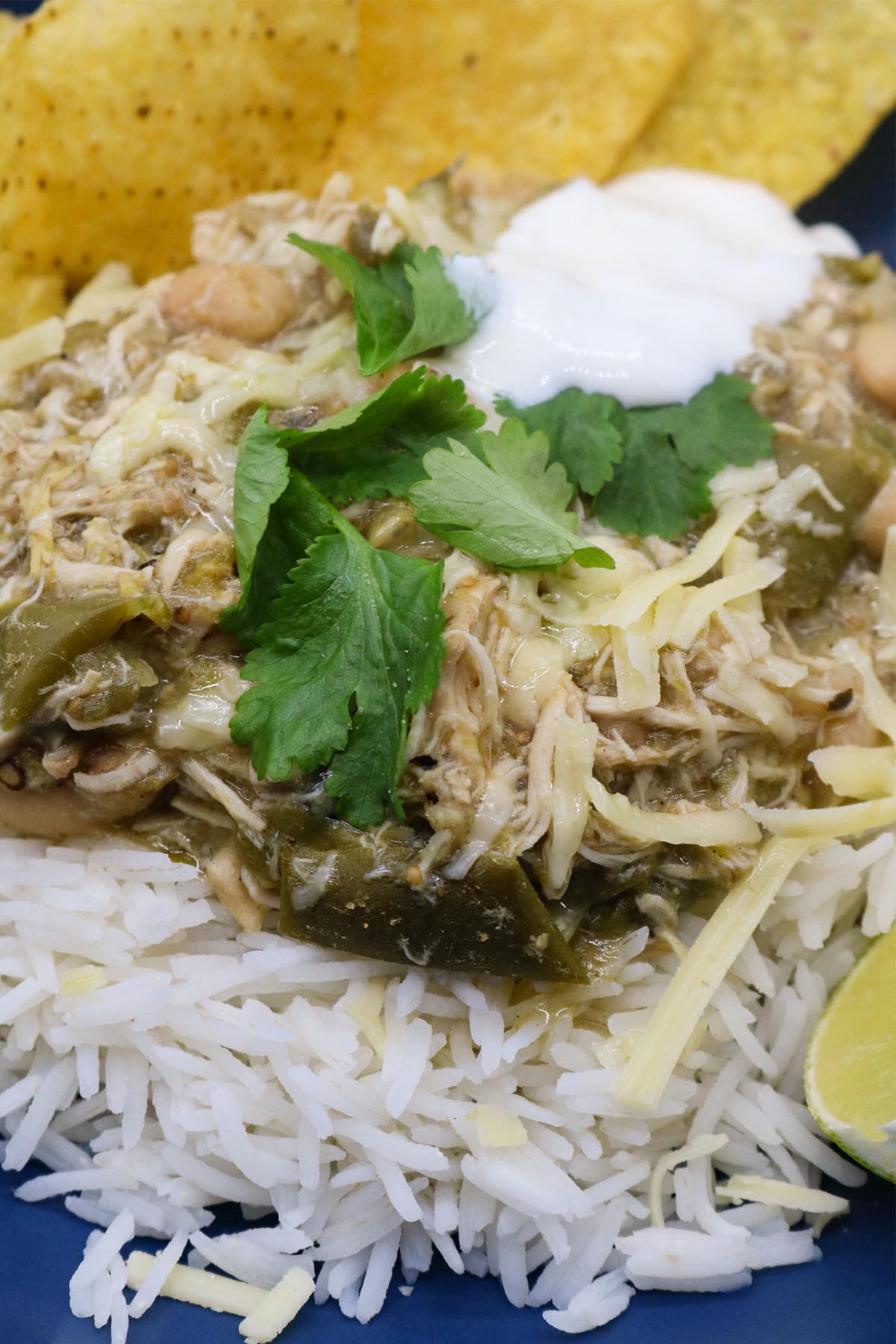 Close up of white chicken chilli in blue bowl with rice, tortilla crisps, soured cream, coriander and lime wedge