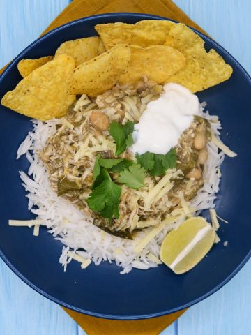White chicken chilli in blue bowl with rice, tortilla crisps, soured cream, coriander and lime wedge