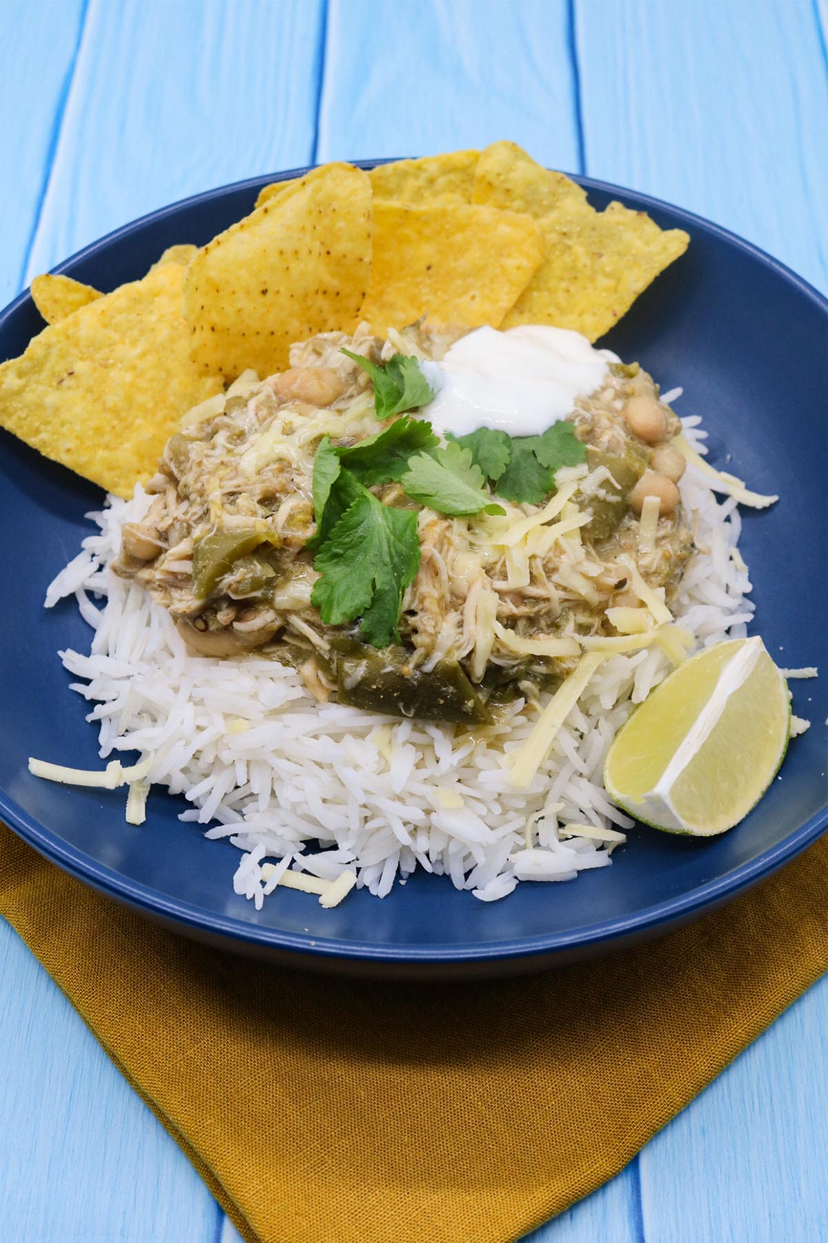 White chicken chilli in blue bowl with rice, tortilla crisps, soured cream, coriander and lime wedge
