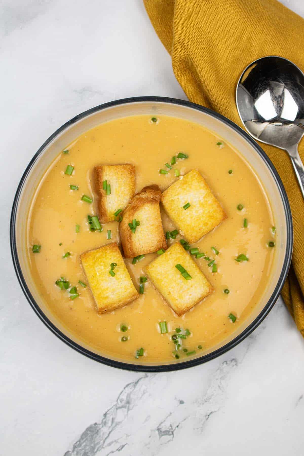 Curried parsnip and sweet potato soup with croutons in a black rimmed bowl with mustard napkin and soup spoon on the side.