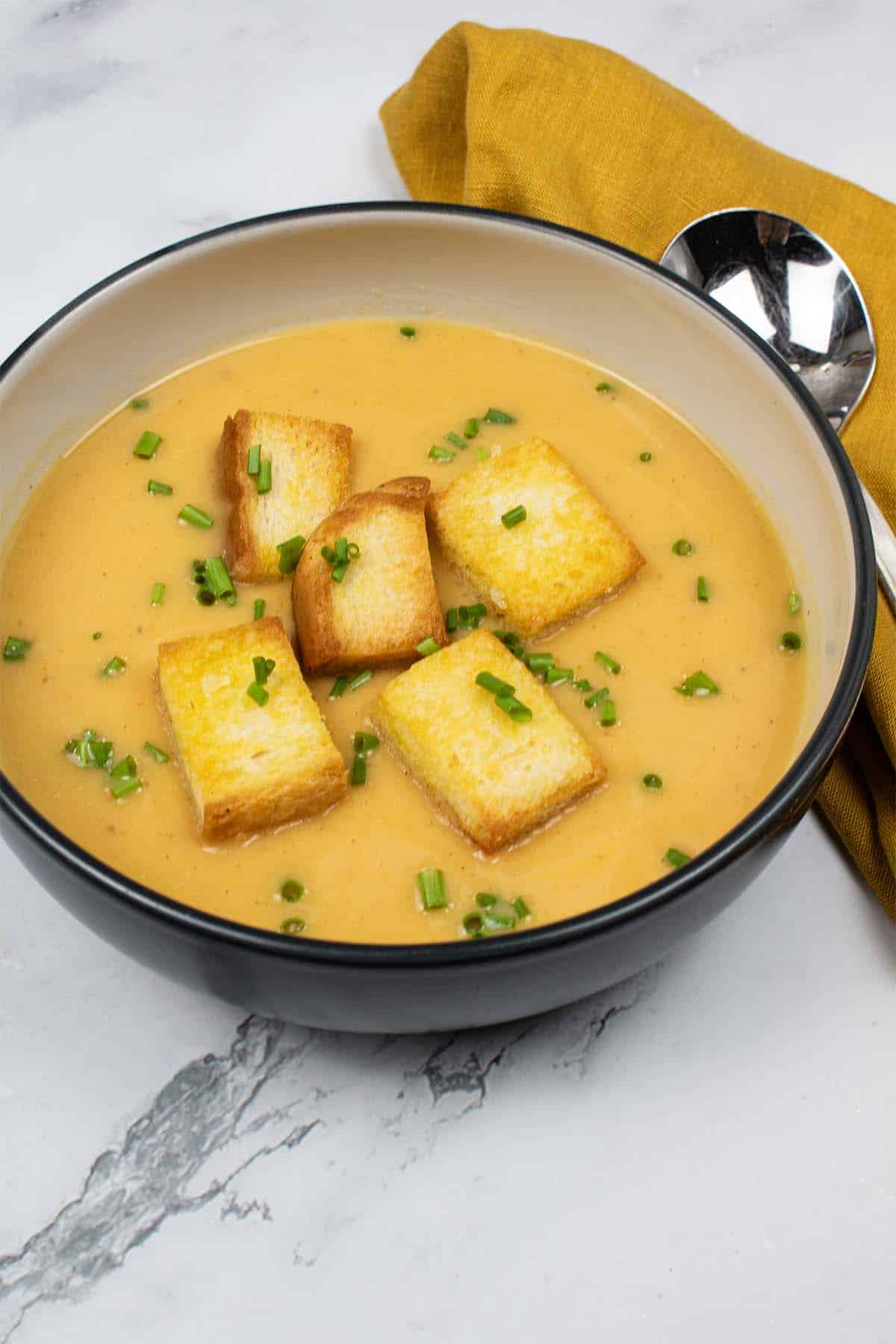 Curried parsnip and sweet potato soup with croutons in a black rimmed bowl with mustard napkin and soup spoon on the side.