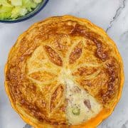 Turkey, gammon and sprout pie in a yellow pie dish with a section of pastry removed. Bowl of cabbage in the background.