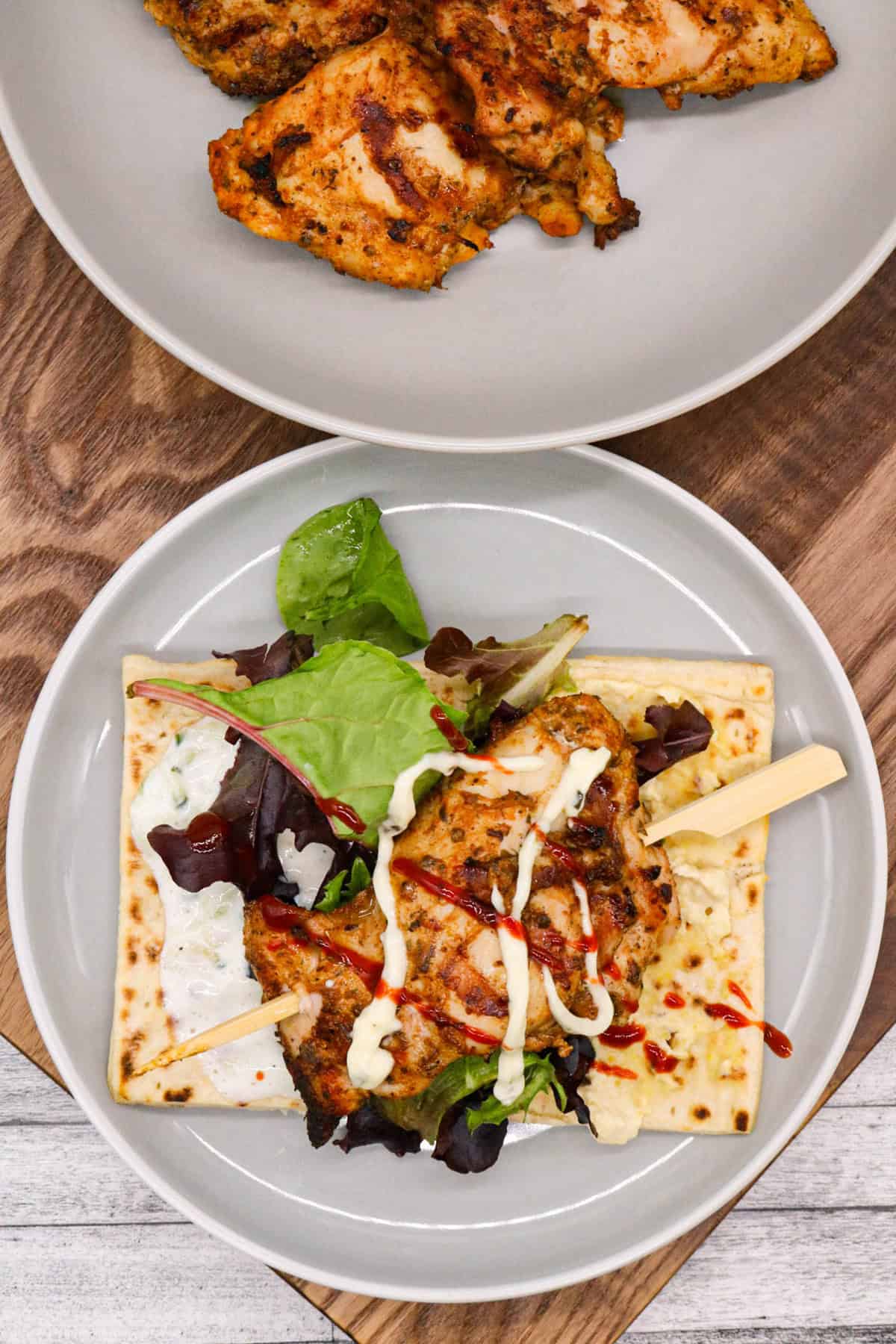 An assembled chicken souvlaki on a wooden skewer, on a flatbread with salad and sauces. A plate of chicken in the background.