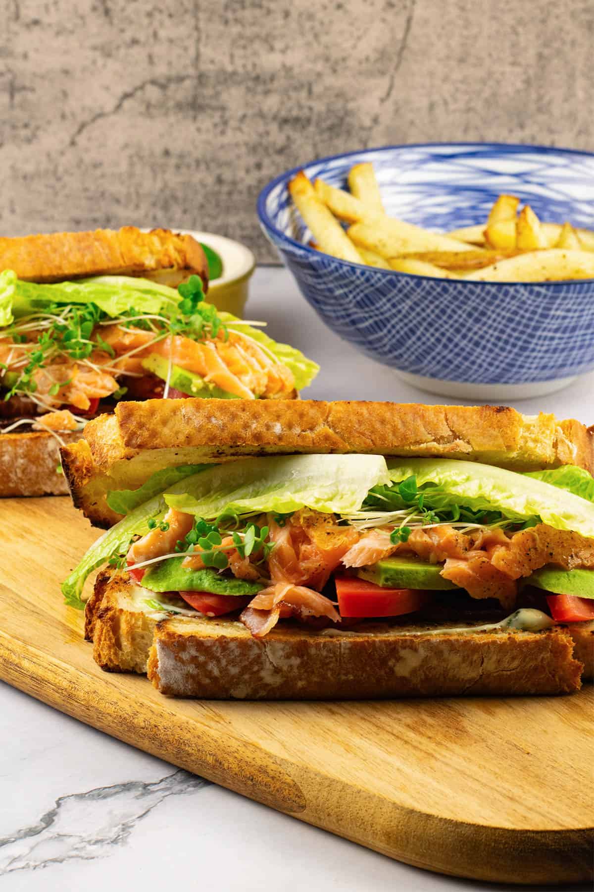2 hot smoked salmon club sandwiches on a board with a bowl of chunky oven chips in the background.