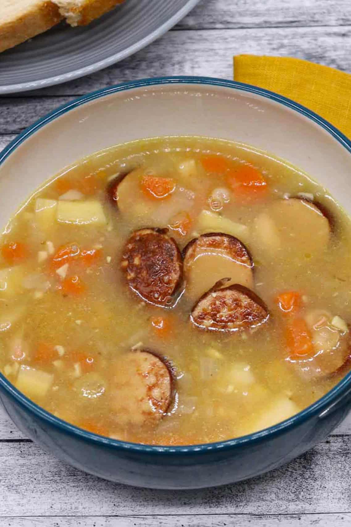 Smoked sausage and bean soup in a bowl with a plate of bread and mustard napkin on the side.