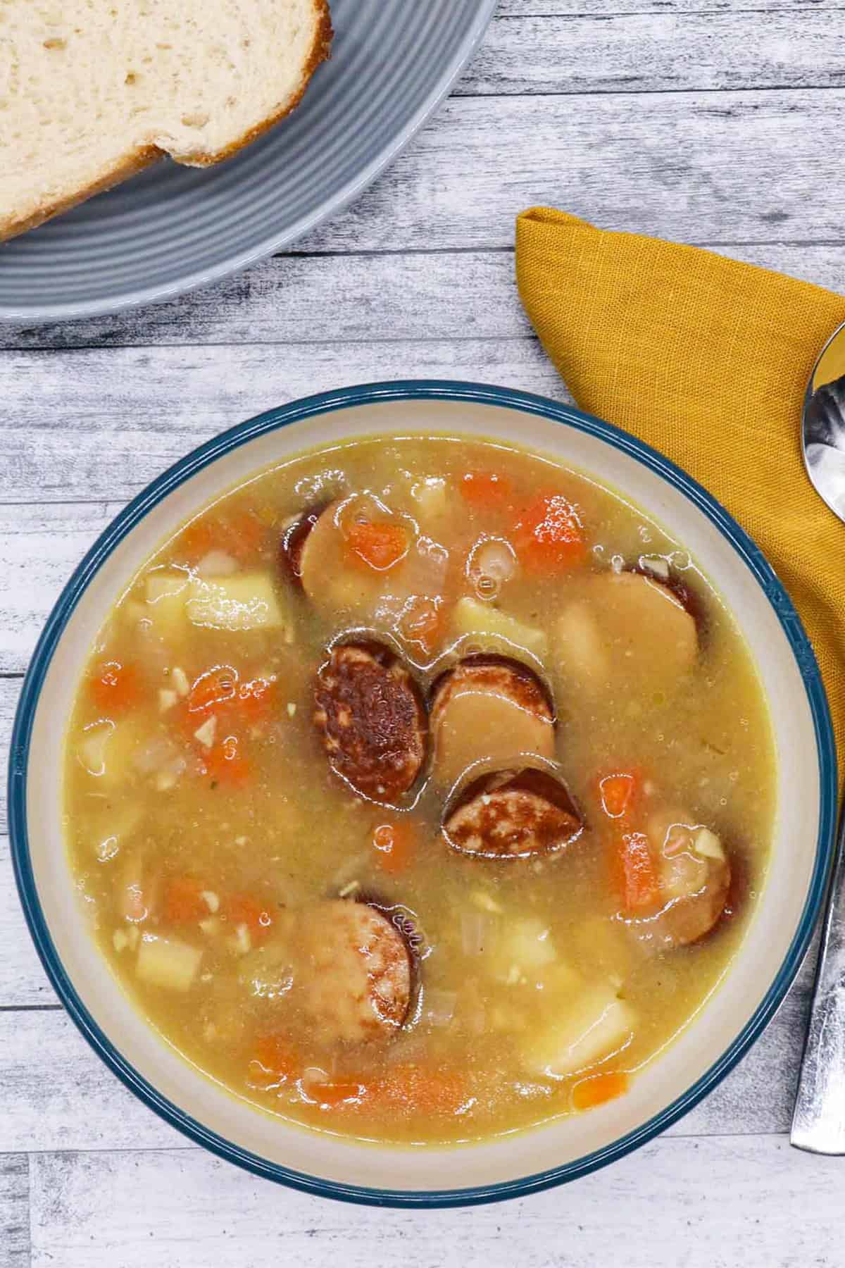 Smoked sausage and bean soup in a bowl with a plate of bread, soup spoon and mustard napkin on the side.
