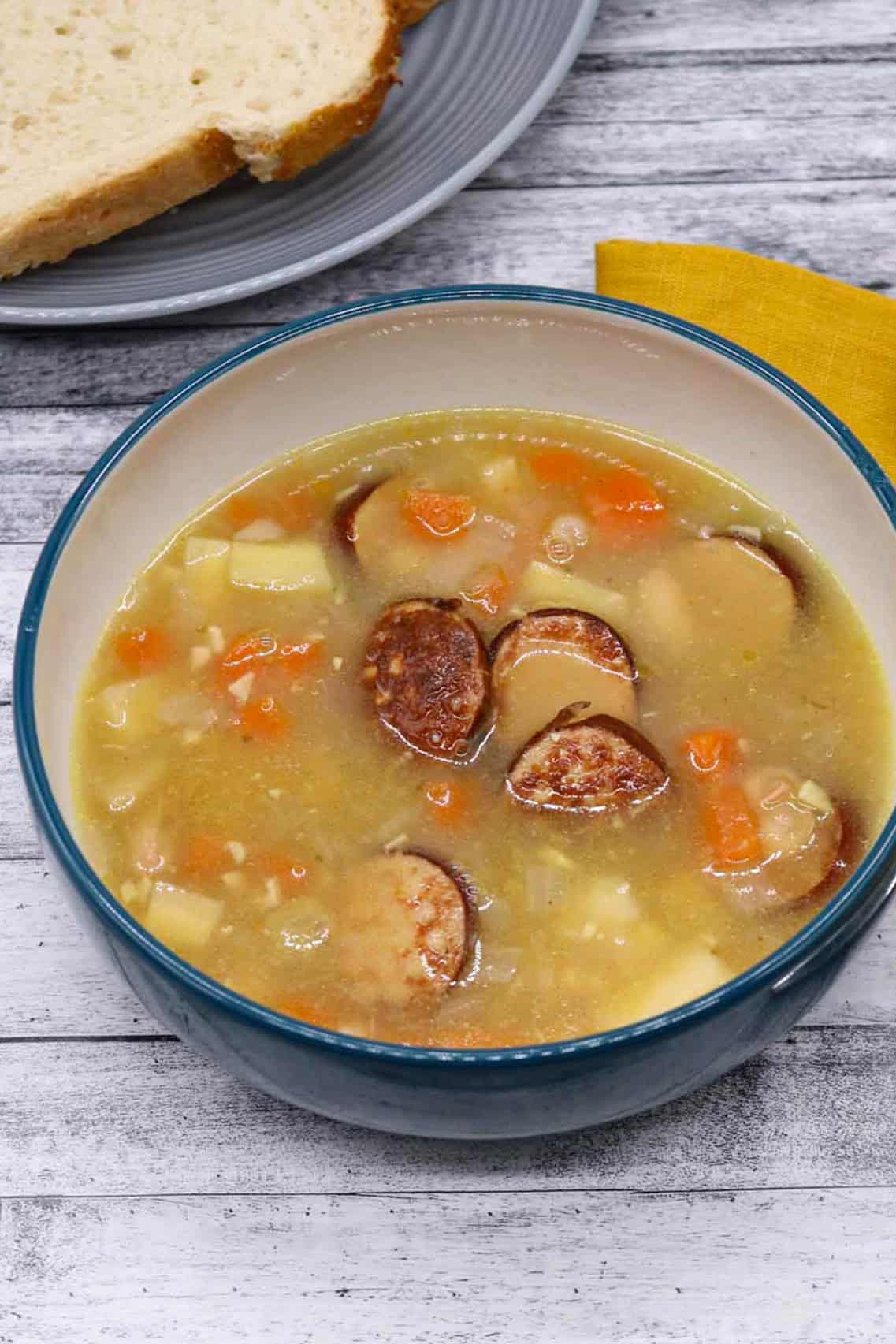 Smoked sausage and bean soup in a bowl with a plate of bread and mustard napkin on the side.