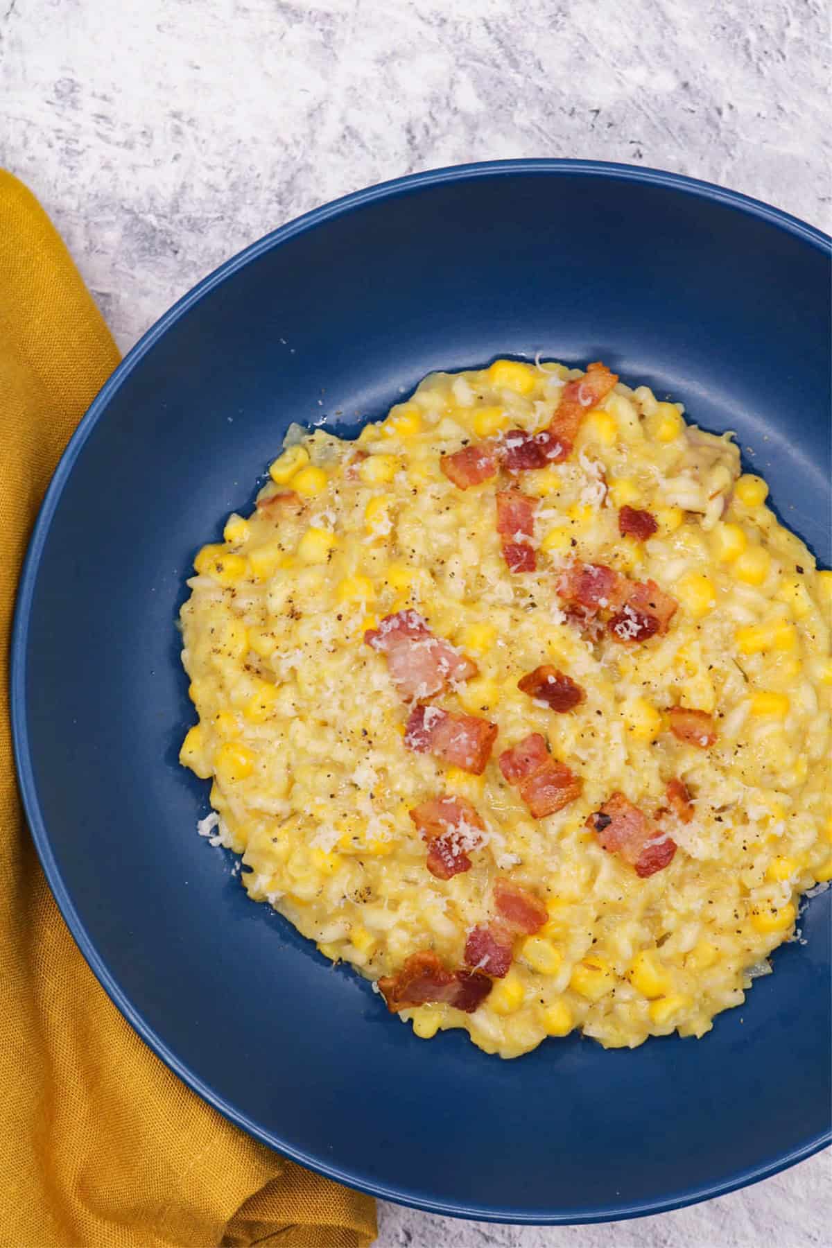 A portion of sweetcorn and bacon risotto in a dark blue bowl with a mustard napkin on the side.