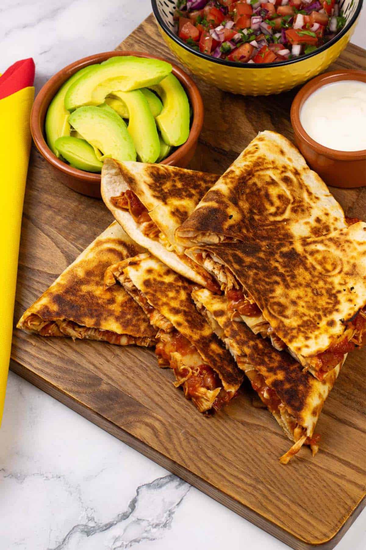 Chicken tinga quesadillas on a board with a bowl of salsa and dishes of sliced avocado and sour cream.