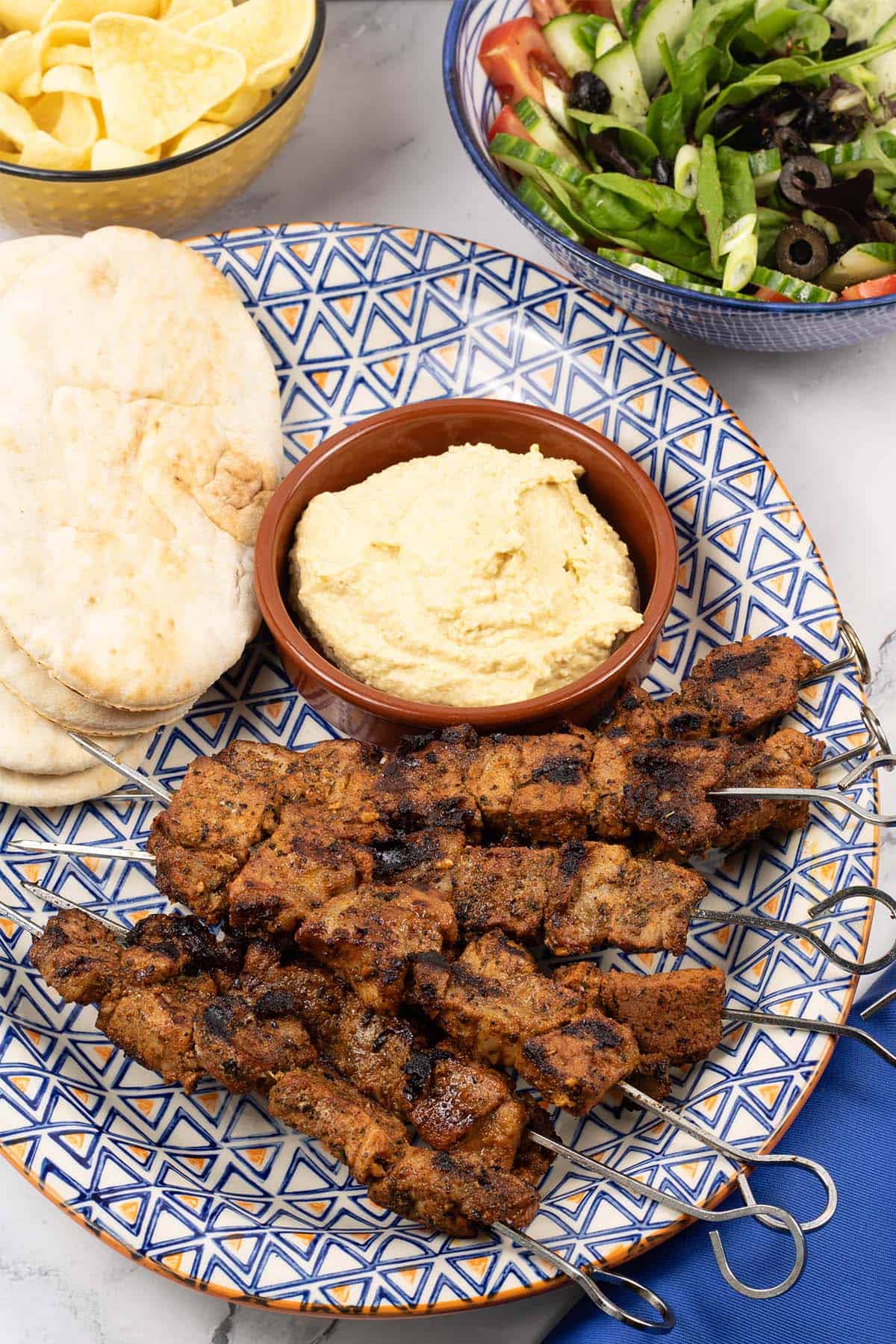 Easy greek pork skewers on an orange blue and white patterned platter with pitta breads and houmous.