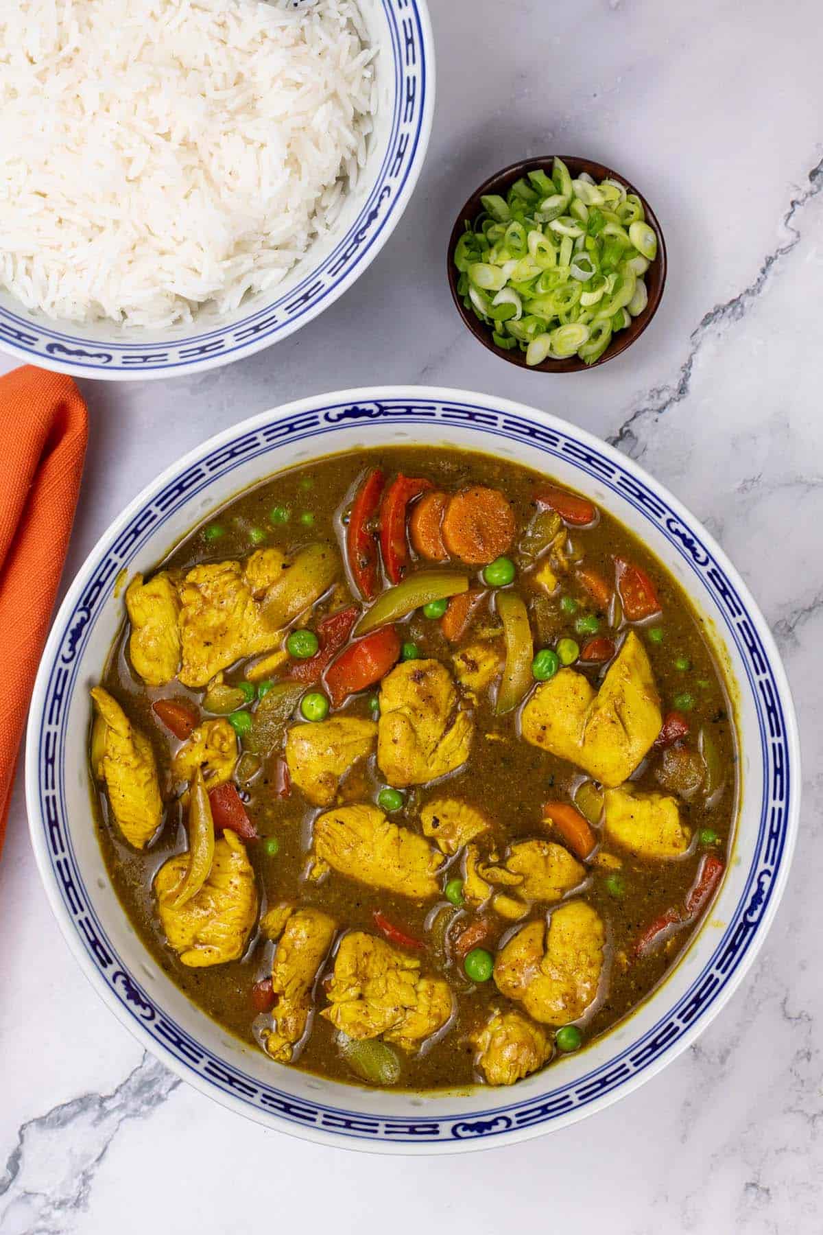 Fakeaway Chinese chicken curry in a blue and white oriental serving bowl with a bowl of rice and dish of sliced spring onions on the side.