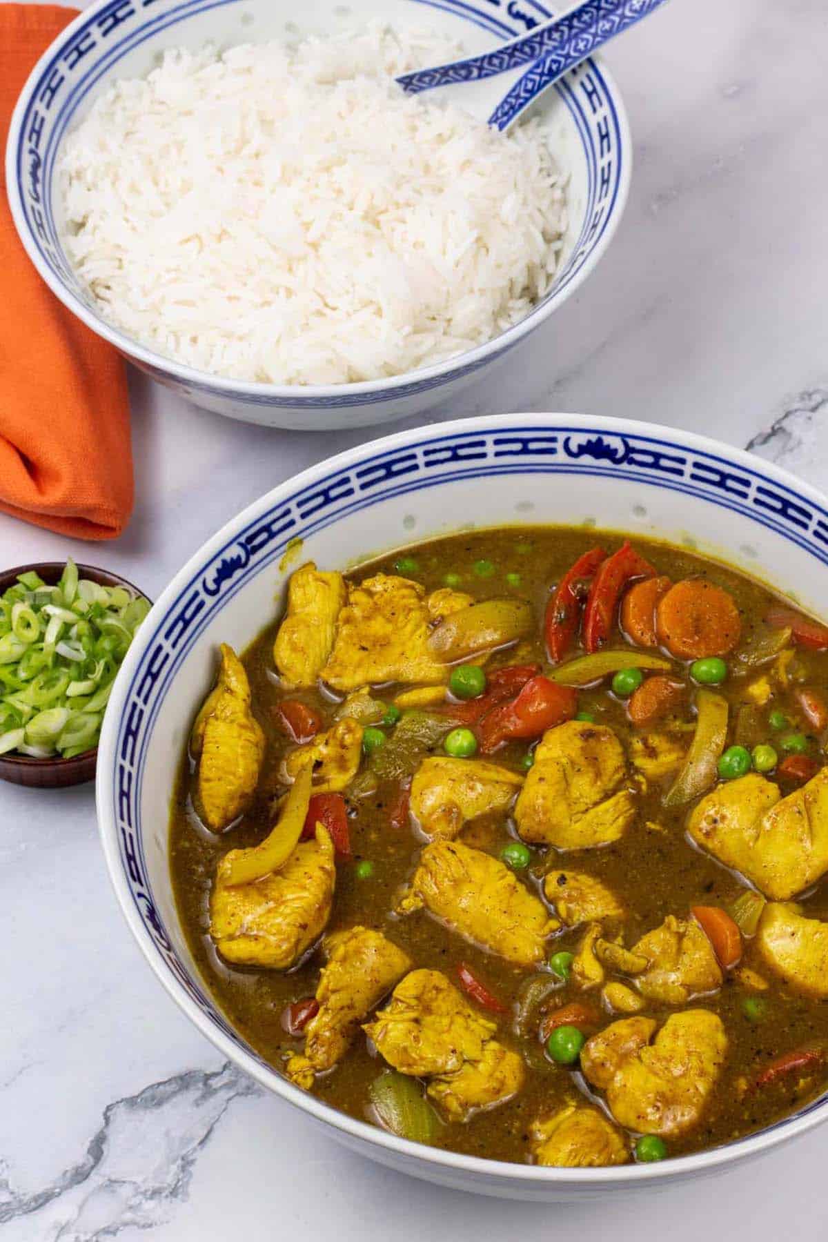 Fakeaway Chinese chicken curry in a blue and white oriental serving bowl with a bowl of rice and dish of sliced spring onions on the side.