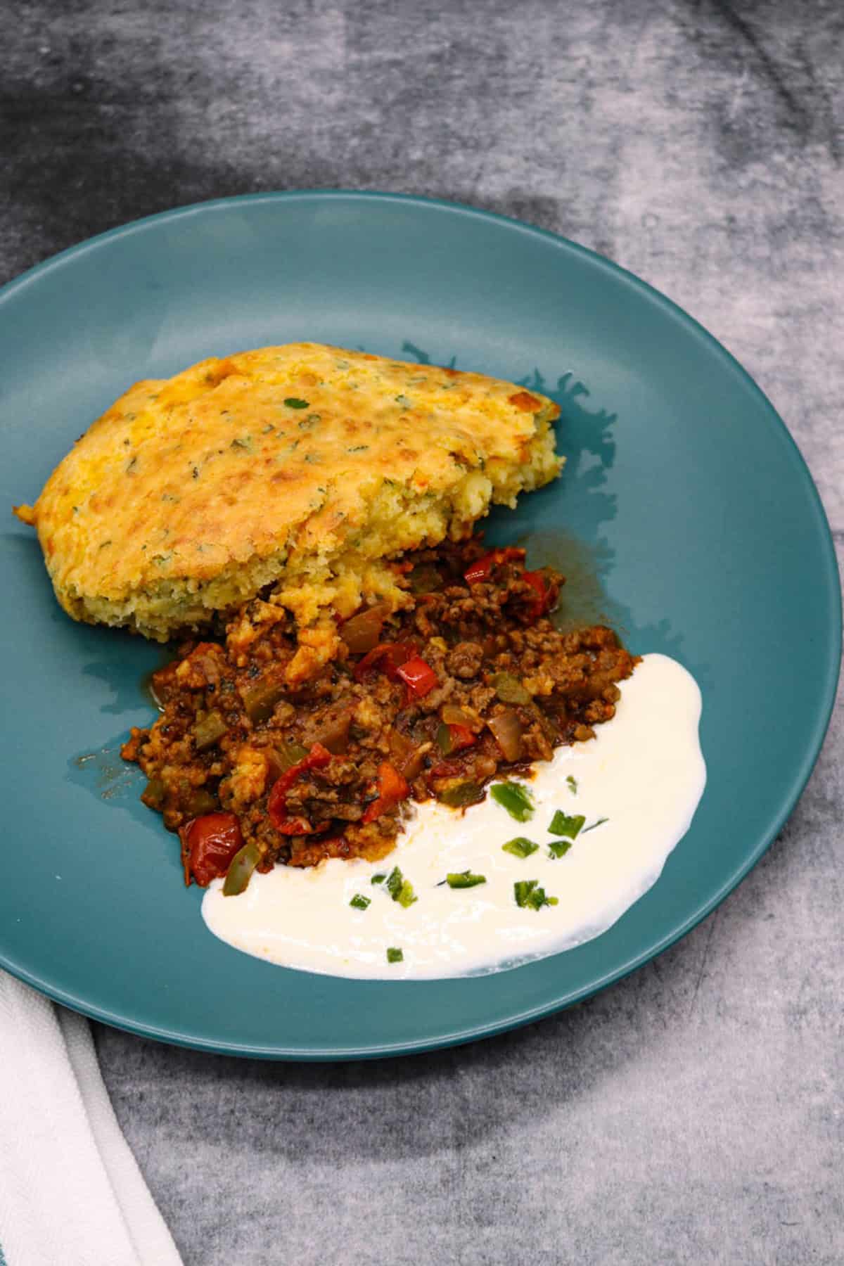 A portion of chilli cornbread pie on a teal dinner plate with some sour cream on the side.
