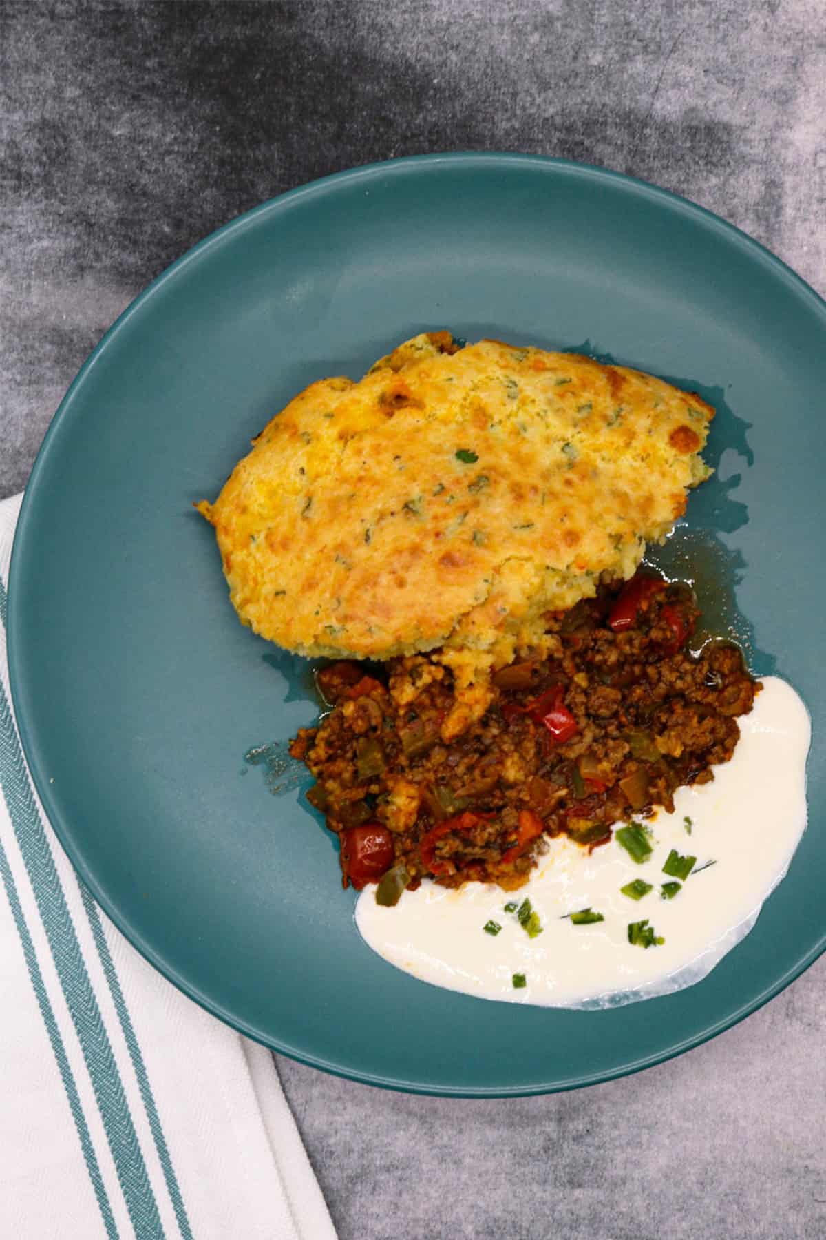 A portion of chilli cornbread pie on a teal dinner plate with some sour cream on the side.