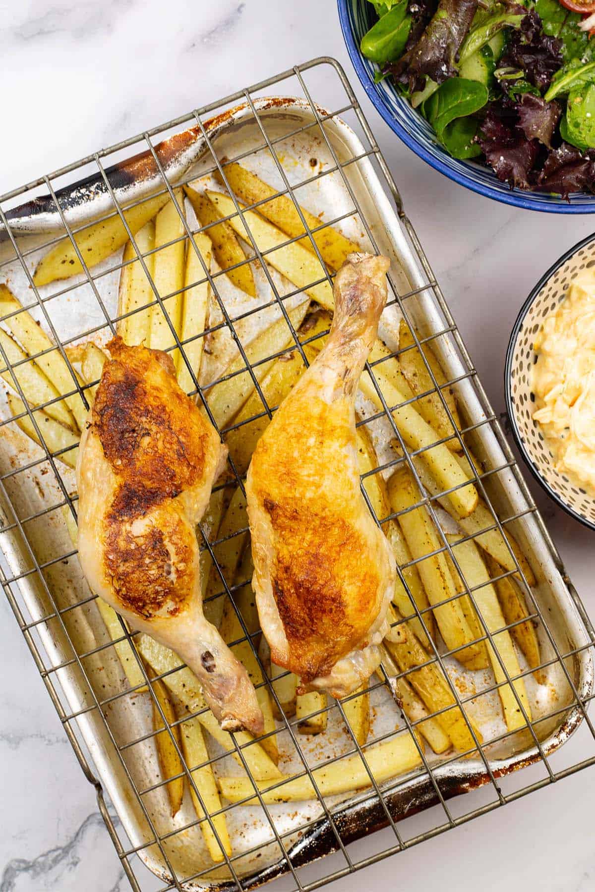 Easy chicken legs and chips traybake in a roasting tin with bowls of coleslaw and salad on the side.