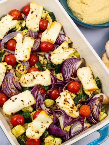 Halloumi and mediterranean veg tray bake in a large rectangle Staub dish, with pitta breads and houmous on the side.