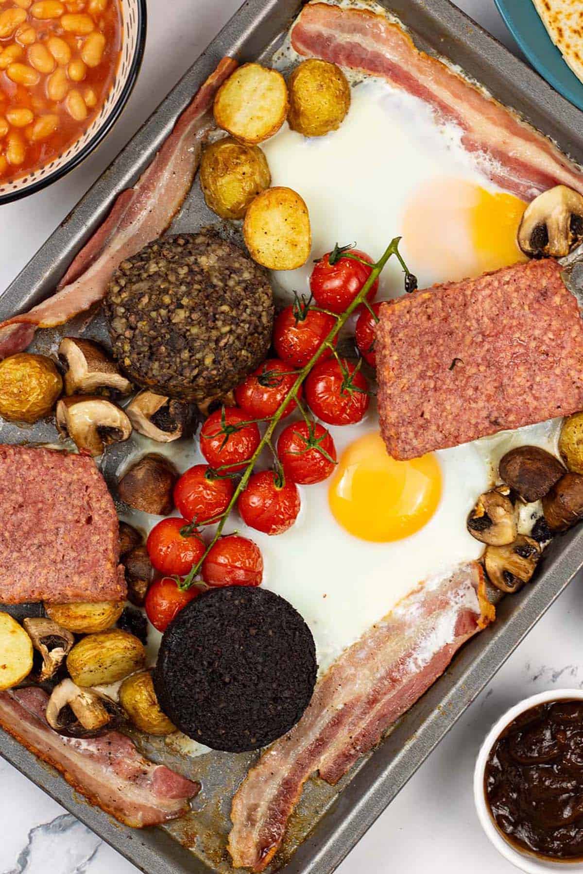 Scottish breakfast traybake on a baking tray with a bowl of baked beans, plate of tattie scones and small dishes of brown and tomato sauces on the side.