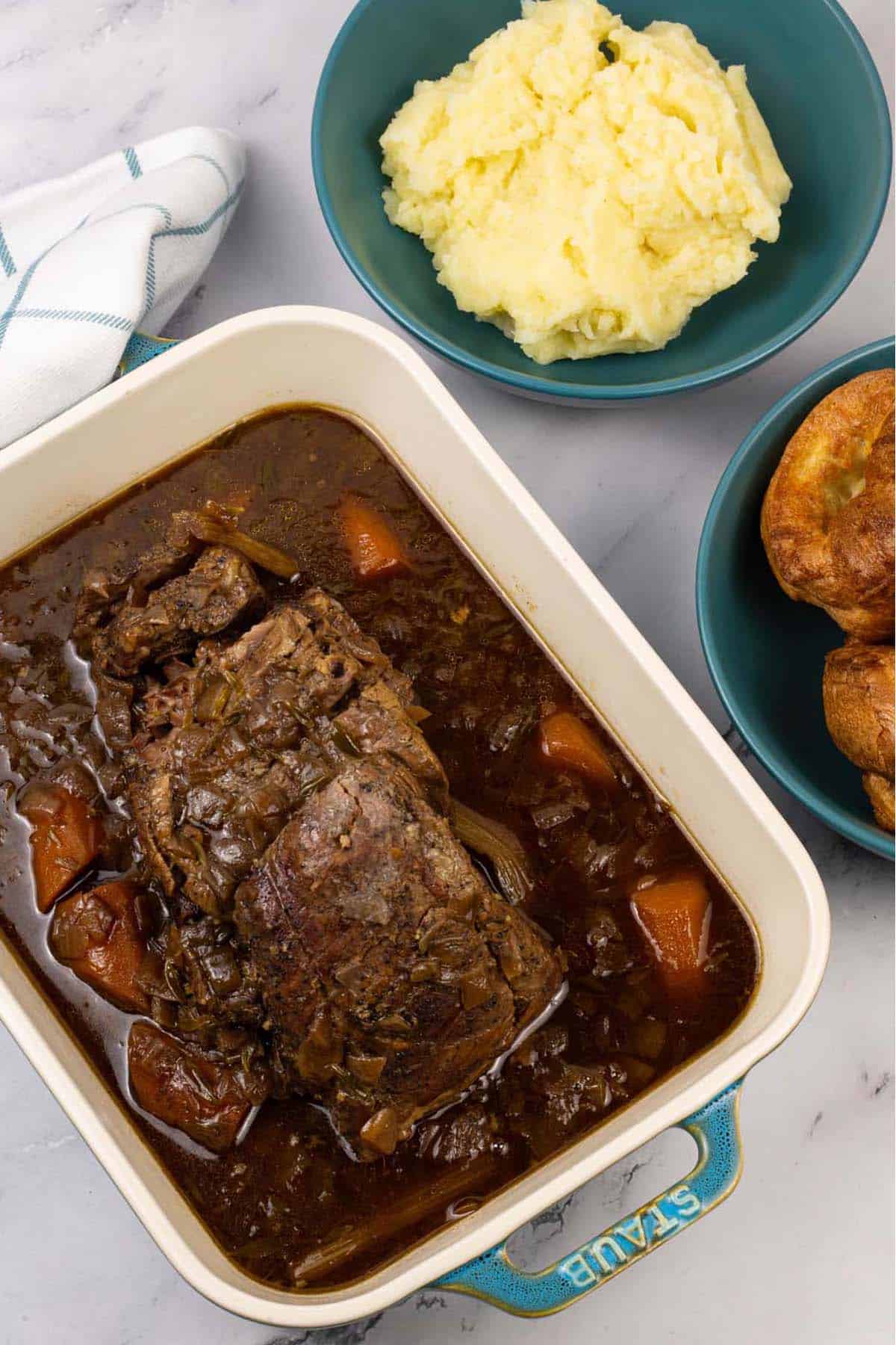 Slow cooked brisket with red wine, thyme and onions in a rectangular serving dish with bowls of mashed potatoes and Yorkshire puddings on the side.
