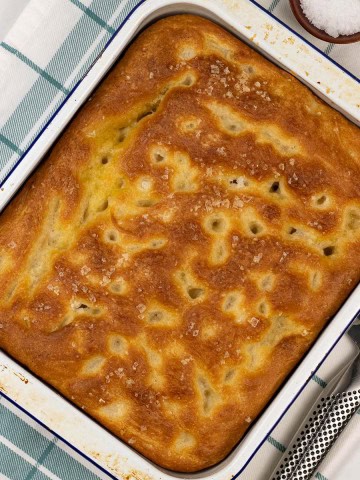 Authentic focaccia in a white and blue rimmed enamel dish, sitting on a teal and white checked towel.