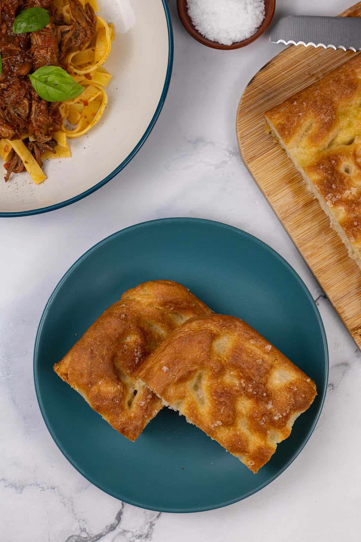 Two slices of authentic focaccia on a teal side plate with the rest on a board and a bowl of slow cooker beef ragu in the background.