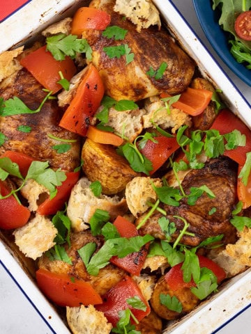 Chicken thighs with romesco traybake in a white enamel roasting tin with a bowl of salad in the background.