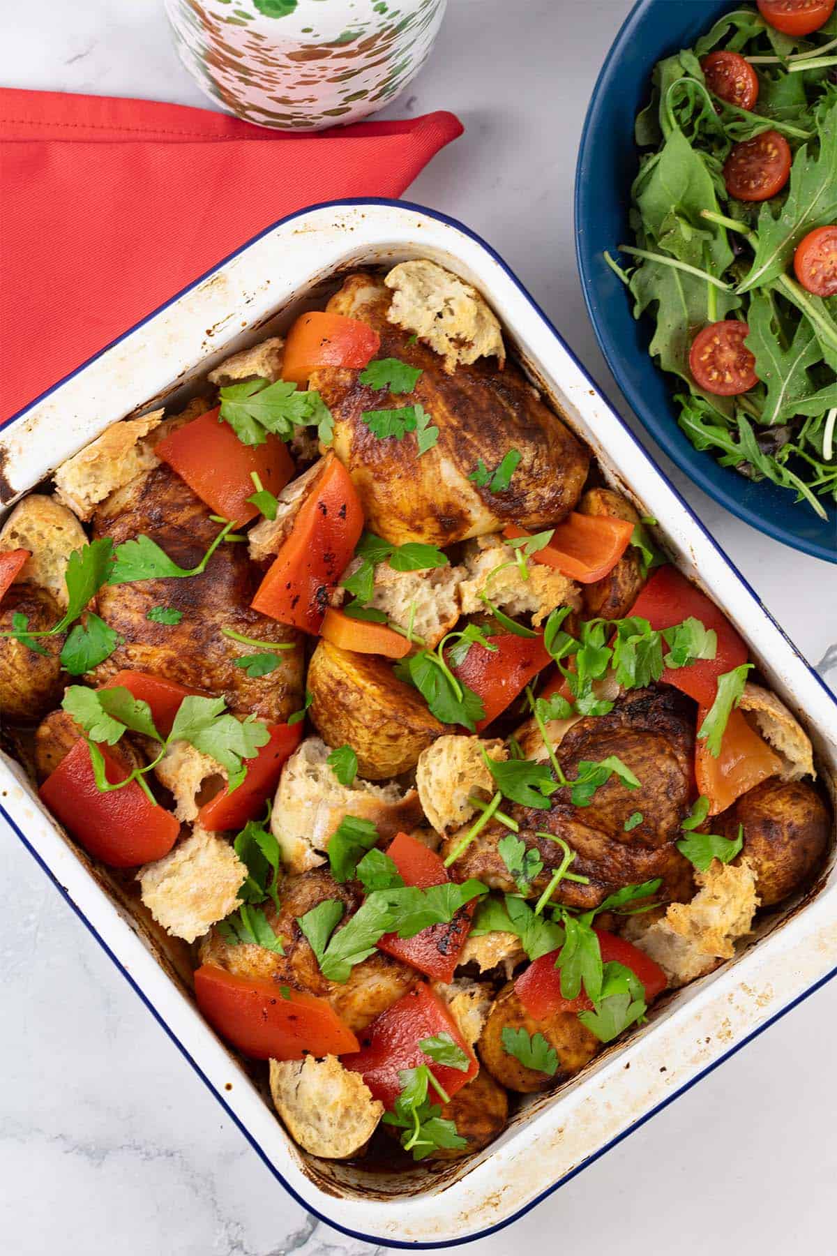 Chicken thighs with romesco traybake in a white enamel roasting tin with a bottle of oil and bowl of salad in the background.