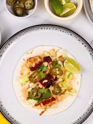Grilled salmon tacos with chipotle lime yogurt on a white and black dinner plate with dishes of chipotle yogurt, tortillas, jalapeños, lettuce and lime wedges in the background.
