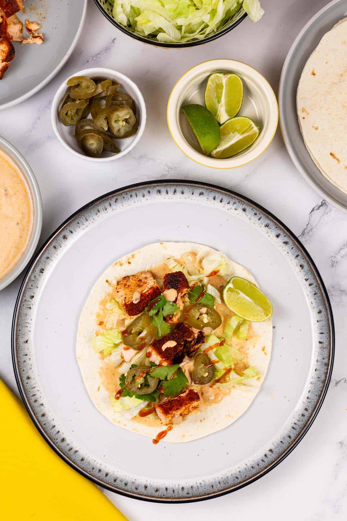 Grilled salmon tacos with chipotle lime yogurt on a white and black dinner plate with dishes of salmon, flour tortillas, jalapeños, lettuce and lime wedges in the background.