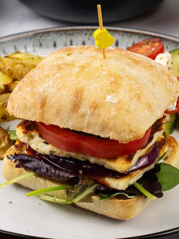 Halloumi burger on a white and black dinner plate with chunky oven chips and greek salad.