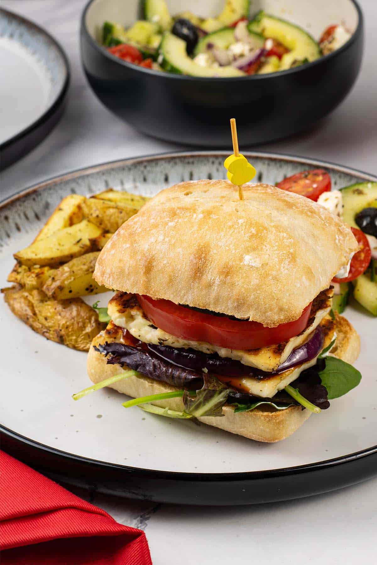 Halloumi burger on a white and black dinner plate with chunky oven chips and greek salad.